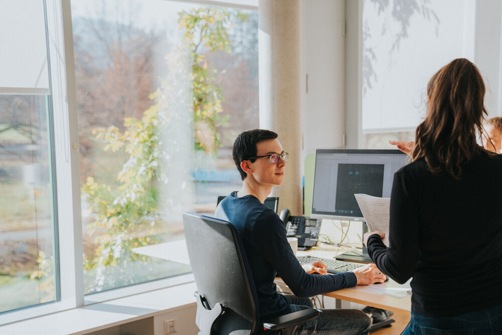 Zwei Personen arbeiten gemeinsam an einem Schreibtisch in einem hellen Büro. Eine Person sitzt vor einem Computerbildschirm, die andere steht daneben und hält ein Blatt Papier. Im Hintergrund sind große Fenster mit Blick ins Grüne.