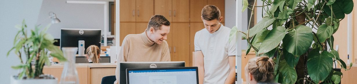 Zwei Personen arbeiten gemeinsam an einem Schreibtisch in einem hellen Büro. Eine Person sitzt vor einem Computerbildschirm.