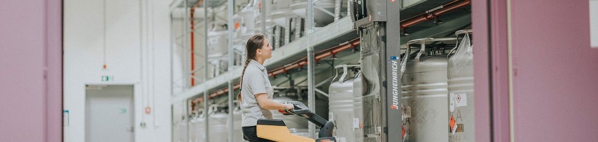 Eine Person bewegt ein elektrisches Hubfahrzeug durch ein Lager mit großen Edelstahlbehältern, die in hohen Regalen gelagert sind