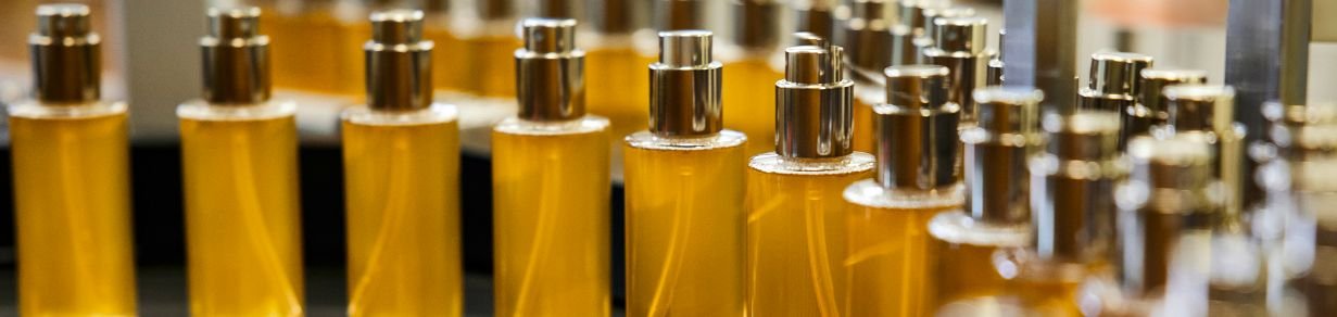 Mehrere Glasflaschen mit goldfarbener Flüssigkeit und silbernen Sprühverschlüssen stehen in Reihen auf einer Produktionsfläche